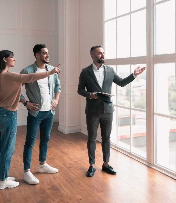 Immobilienmakler zeigt einem lächelnden Paar eine lichtdurchflutete Wohnung mit großen Fenstern.